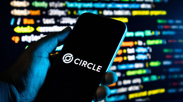 Hand holding a smartphone with the circle logo on the screen against a background of computer code