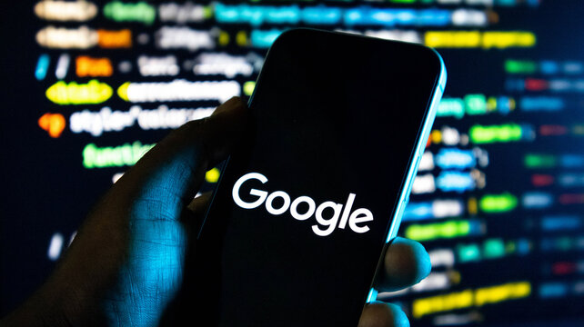 Closeup of a hand holding a smartphone displaying the google logo against a background of blurred computer code