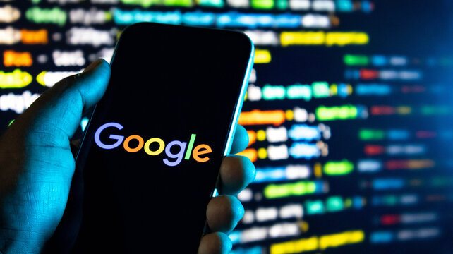Hand holding a smartphone displaying the google logo with a blurred background of computer code on a screen