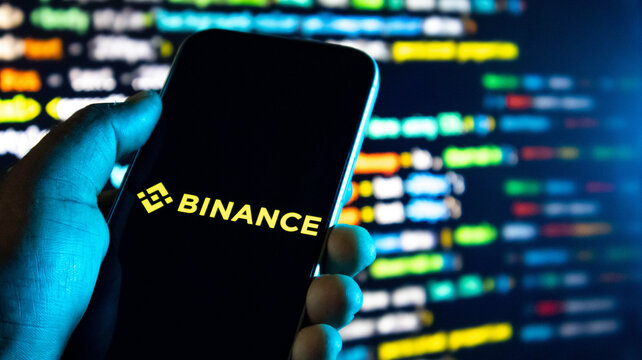 Hand holding a smartphone with the binance cryptocurrency exchange logo on the screen against a background of computer code