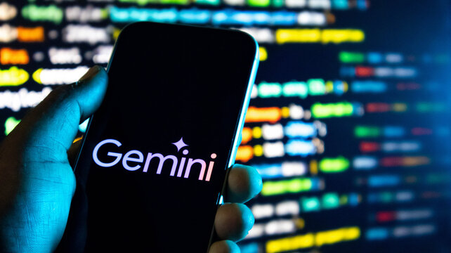 Closeup of a hand holding a smartphone displaying the google gemini ai logo against a background of blurred computer code