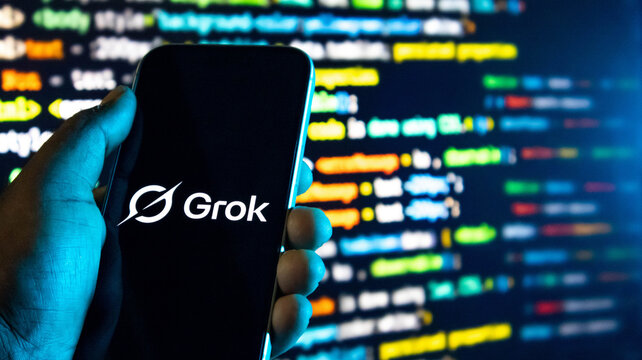 Hand holding a smartphone displaying the grok ai logo against a background of blurred computer programming code