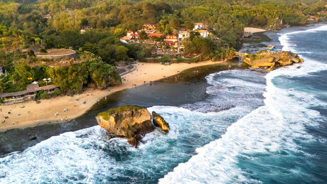 Aerial view of Mesra Beach with golden sand, turquoise waves crashing against rocky outcrops, and lush green vegetation in Kecamatan Tanjungsari, Daerah Istimewa Yogya, Indonesia.