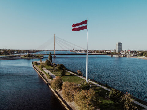 Aerial view of the Latvian flag flying on a tall flagpole in a park by the Daugava River, with the Vansu Bridge in the background in Riga, Latvia.