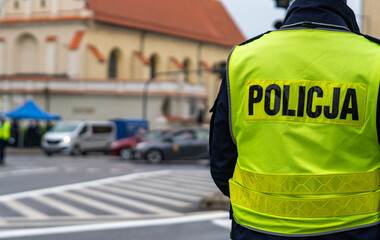 Back view of a Polish police officer wearing a bright, high-visibility yellow vest with the word POLICJA, standing at a city intersection near a pedestrian crossing, with blurred urban street backgrou © Adam