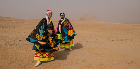 Fototapeta premium Two young Egyptian folk dancers wearing vibrant multicolored embroidered robes and turbans pose proudly before the Pyramids of Giza across the hazy