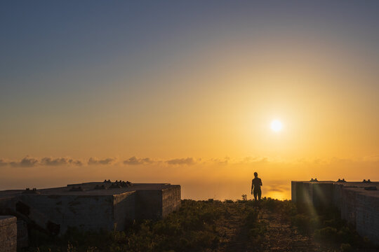 Silhouette of a woman walking towards a golden sunrise past old concrete blocks on the summit of Mount Edheri in Crete, Greece