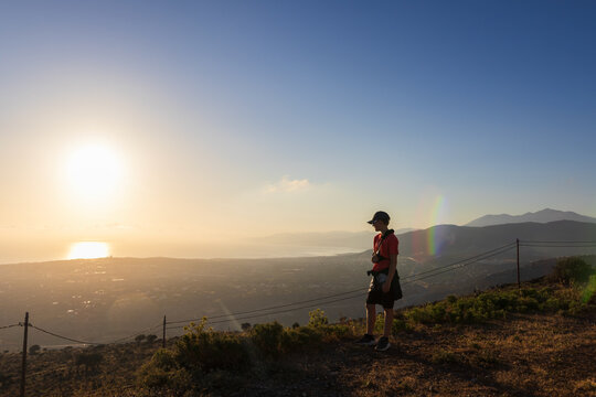 An 11-year-old boy with an arm brace stands on the top of Mount Edheri at sunset, Crete coast. Inspiring travel concept of physical limits, pediatric injury recovery, and youth hiking adventure