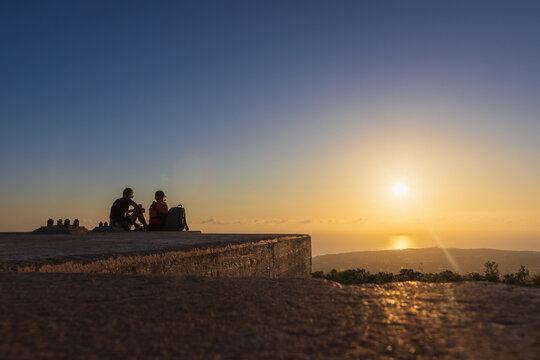 Silhouette of a woman and her eleven-year-old son with a backpack sitting on a concrete block and watching the sunrise together on Mount Edheri in Crete, Greece