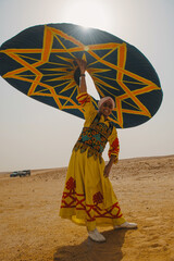 Fototapeta premium Joyful folk dancer in embroidered yellow traditional costume raises a bold teal and orange star patterned ceremonial umbrella overhead in a vast sunny