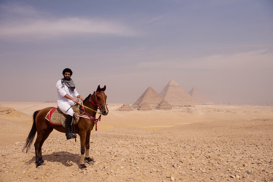 Confident Arab horseman wearing keffiyeh rides brown horse on sandy Giza plateau with iconic Egyptian pyramids rising in background under hazy sky