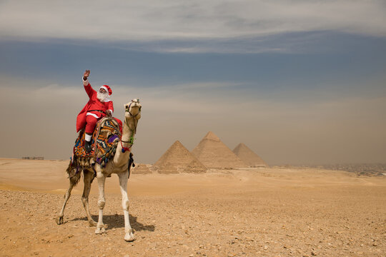 Joyful Santa Claus riding a colorfully decorated camel and waving high before the three great Giza pyramids across a sunlit Egyptian desert plateau