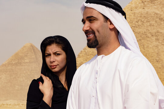 Authentic Arab couple in abaya and thobe showing playful tension with skeptical woman glancing sideways at smiling man before softly blurred Giza