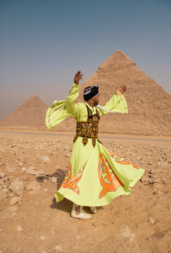 Whirling dancer in embroidered lime and orange tanoura costume spins with raised arms before iconic Giza pyramids in rocky desert