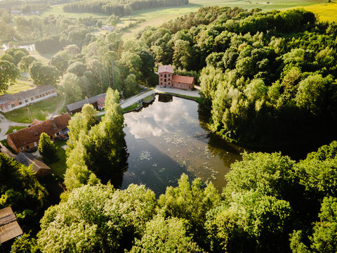 Aerial view of a historic red-brick building complex situated beside a calm pond surrounded by dense green forests and lush trees in the countryside of Poland.