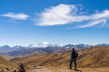 A hiker stands on a ridge near Palccoyo Rainbow Mountain, looking at the snow-capped peak of Mount Ausangate under a bright blue sky, Peru © John N