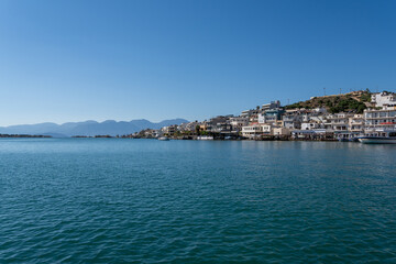 Fototapeta premium Waterfront buildings of Elounda coastal town line Mirabello Bay shoreline on Crete island under sunny blue sky with mountains.