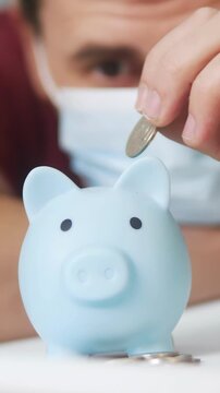 A blue piggy bank is placed on a table as a man leans in closer, observing the piggy bank. The man, wearing a mask, focuses intently on the piggy bank.