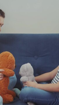 A woman and a girl sit together on a couch, each holding a stuffed toy. The woman holds a brown teddy bear while the girl holds a gray stuffed bunny.
