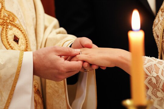 Priest places wedding ring on bride finger during church ceremony. Couple exchanges rings, symbolizing love and commitment. Candle flame burns brightly, highlighting sacred union, holy vows.