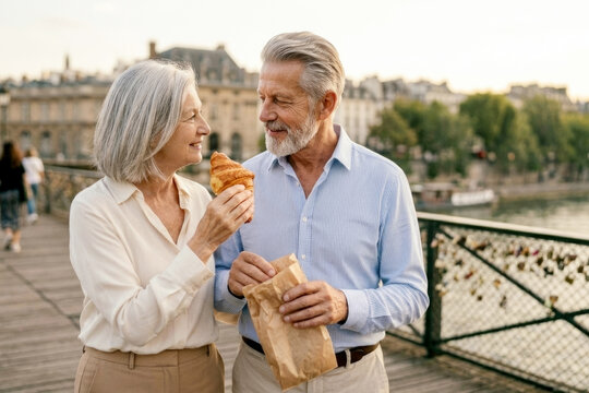 Happy senior couple sharing a fresh croissant on bridge representing authentic retirement romance