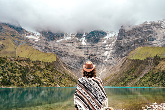 Rear view of a woman in traditional poncho and hat looking at the blue lake of Humantay in Peru