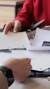 Hands review white printed sheets at office table, with red sleeve and wristwatch visible while papers are sorted, passed and adjusted over stacked documents during meeting discussion