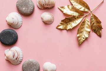 Obraz premium Sea shells and smooth stones arranged on pastel pink surface with golden leaves creating a decorative border. Empty space for text at center.