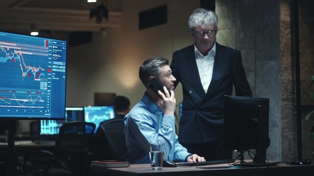 Caucasian male broker talking on smartphone while working with financial charts on computer. Senior businessman standing beside trader in dark office. Team monitoring stock market data at night.