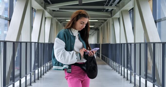 Student texting on glass bridge with varsity jacket and tote, structural beams and long railings frame the scene, checking map and messages while planning route, calm campus atmosphere