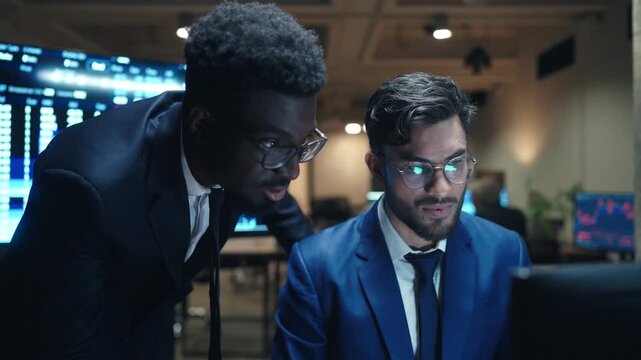 Diverse male brokers wearing glasses working at computer in dark trading office. One trader leaning toward colleague while analyzing financial charts. Team discussing stock market data at night.