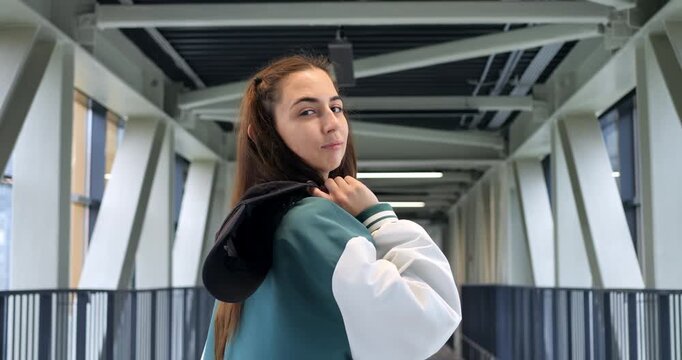 Young woman posing with jacket over shoulder in bright corridor, playful glance and subtle smile, fanny pouch and varsity jacket contrast with industrial beams, confident casual model vibe