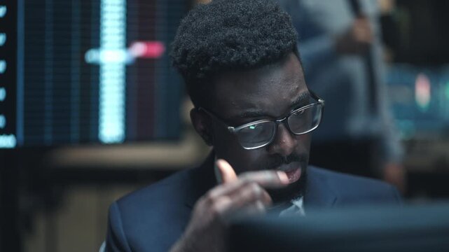 Close up African American male broker wearing glasses and looking at computer screen in dark trading office. Trader analyzing stock market data with financial charts around him late at night.