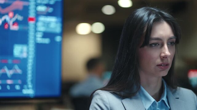 Serious Caucasian female broker looking at computer screen near financial chart monitor. Woman trader analyzing stock market data in dark office. Professional focusing on risky investments at night.