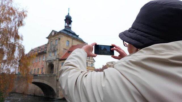 Female tourist with a mobile phone capturing the iconic altes rathaus, the old town hall, over the regnitz river in bamberg, germany during a cloudy autumn day revealing the landmark