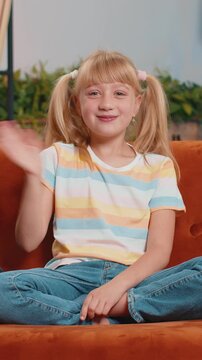 Young blonde child kid lady smiling friendly at camera and waving hands gesturing hello, hi, greeting or goodbye, welcoming with hospitable expression. Teen toddler at home in living room sits on sofa