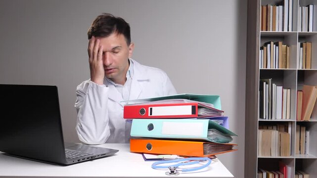 Overworked male doctor slumped at a cluttered desk with laptop and towering patient folders, exhausted and stressed by paperwork, long hours and administrative burden in a clinical office