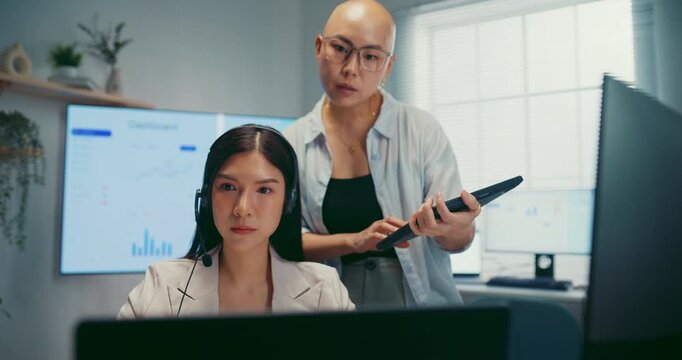 Young Asian businesswoman hold tablet to mentor trainee in call center workspace office. Corporate customer service training, employee support coach, telemarketing agent, teamwork help desk concept.