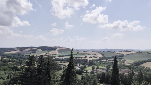 Panorama collinare visto da Urbisaglia, con alberi in primo piano, campi coltivati, boschi e colline che si estendono fino all&rsquo;orizzonte sotto cielo azzurro con nuvole leggere