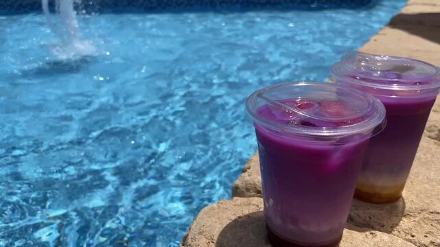 Purple and orange gradient fruit drinks with lids resting poolside as water splashes from a fountain in bright sunlight
