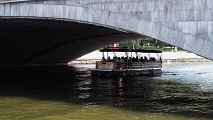 A tranquil moment as a pontoon boat glides smoothly under a concrete bridge in an urban river setting, viewed from below the bridge looking up at the calm and leisurely waterway with brown. © bisonov
