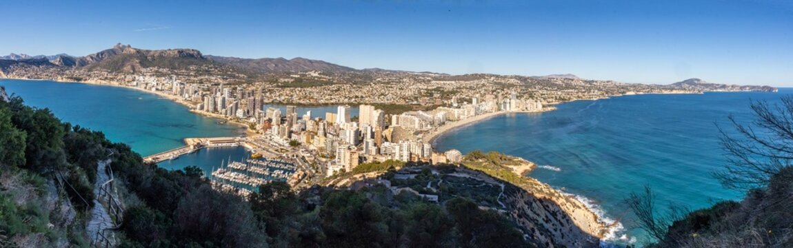 Panoramic view of Calpe, Alicante, from the ascent to Pe&ntilde;&oacute;n de Ifach, overlooking the Mediterranean coastline, marina, sandy beaches, and the surrounding mountains under a clear blue sky.