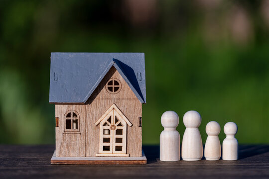 Small miniature house model with a grey roof stands beside a wooden peg doll family on desk with blurred green nature background representing real estate and safety, closeup, outdoors