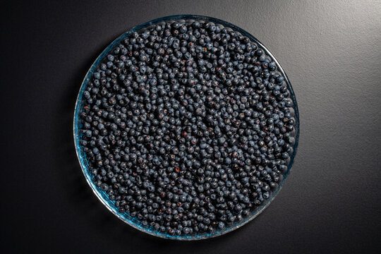 Top view of round glass platter filled with abundance of ripe organic wild blueberries on dark background representing summer harvest, healthy eating and sweet natural dessert ingredients