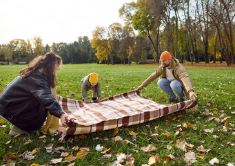 Fototapeta premium Autumn family play with leaves in park. In nature, a happy child helps parents spread a plaid picnic blanket on grass, enjoying outdoors under yellow trees. Celebrates joyful autumn family time.
