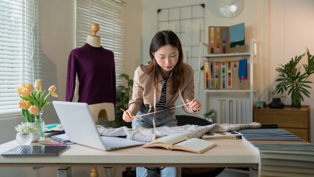 Young Asian fashion designer measuring knitted sweater garment at workspace desk with laptop and fabric samples