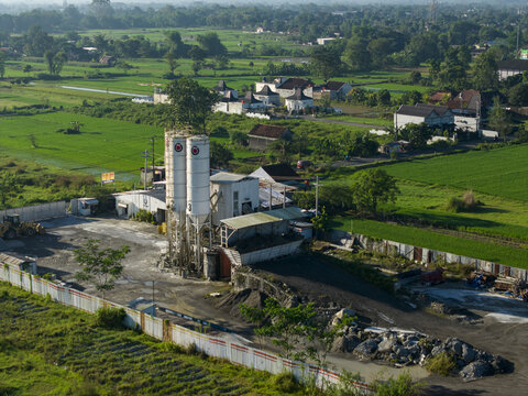 Aerial view of a concrete production site with tall silos, industrial machinery, and piles of aggregate, surrounded by green rice fields in Kecamatan Prambanan, Jawa Tengah, Indonesia.