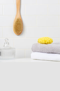 Minimalist bathroom product mockup background with copy space in foreground. Hanging wooden body brush, yellow sea sponge, glass jar, and stacked towels against white subway tile wall.