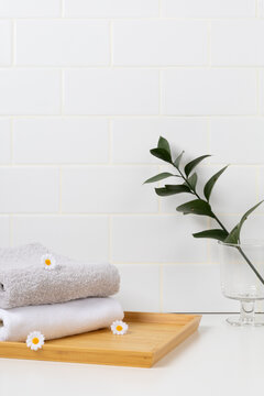 Minimalist spa product mockup background with white subway tile wall. Stacked soft towels on a wooden tray decorated with small daisy flowers and a green leaf branch in glass.