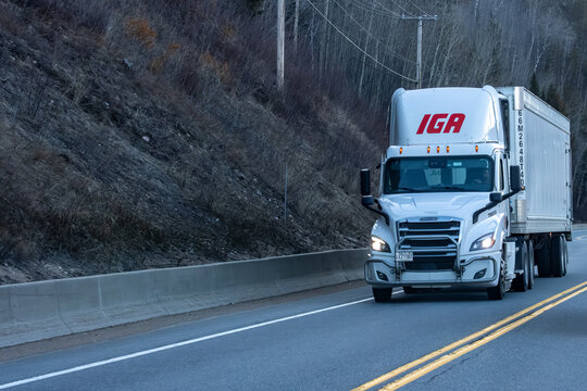 Quebec, Canada, 24 April 2026 : A white IGA Freightliner semi truck drives on a highway, transporting goods along a barren hillside.
